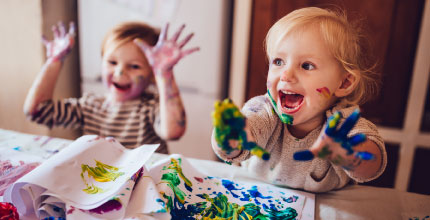 Two toddlers playing with finger paint and smiling. They have paint all over their hands and faces.